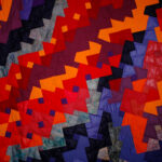 Colorful patchwork quilt with red, orange, purple, and blue geometric patches arranged in diagonal bands.