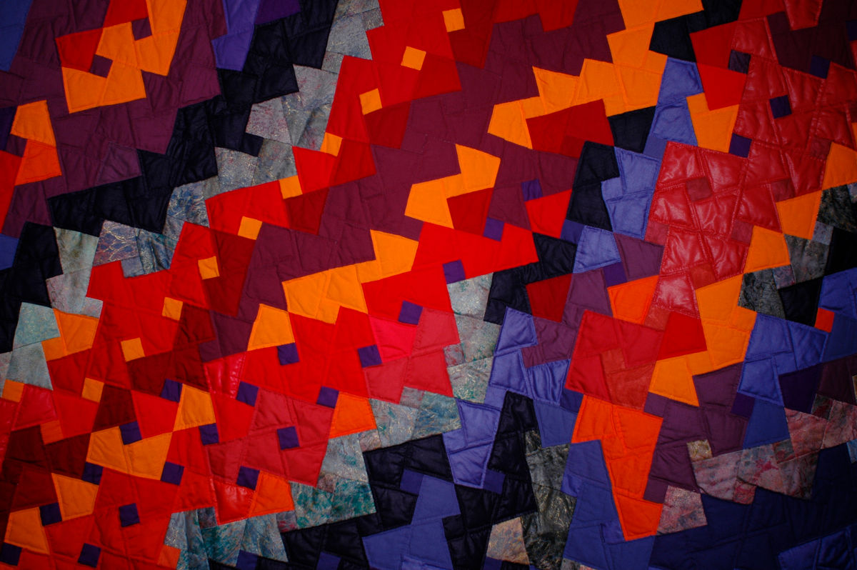 Colorful patchwork quilt with red, orange, purple, and blue geometric patches arranged in diagonal bands.