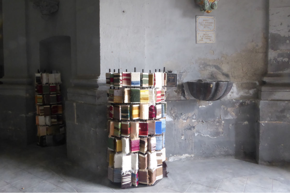 Tall rack of colorful woven fabric swatches in a dim stone interior with a fountain on the wall nearby, suggesting a historic building.