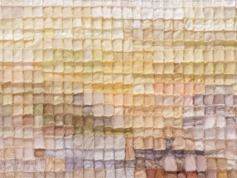 Close-up of a pastel patchwork quilt made of small square fabric patches in pale yellows, creams, and pinks, with visible stitching.