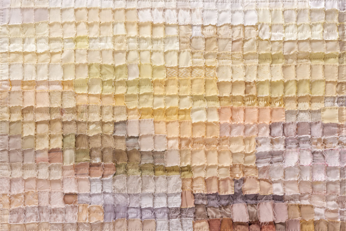 Close-up of a pastel patchwork quilt made of small square fabric patches in pale yellows, creams, and pinks, with visible stitching.