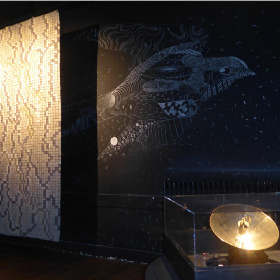 Dimly lit museum exhibit with a tall vertical mosaic wall on the left, a dark mural of a stylized sea creature in the center, and a glass display case containing a glowing brass disk.