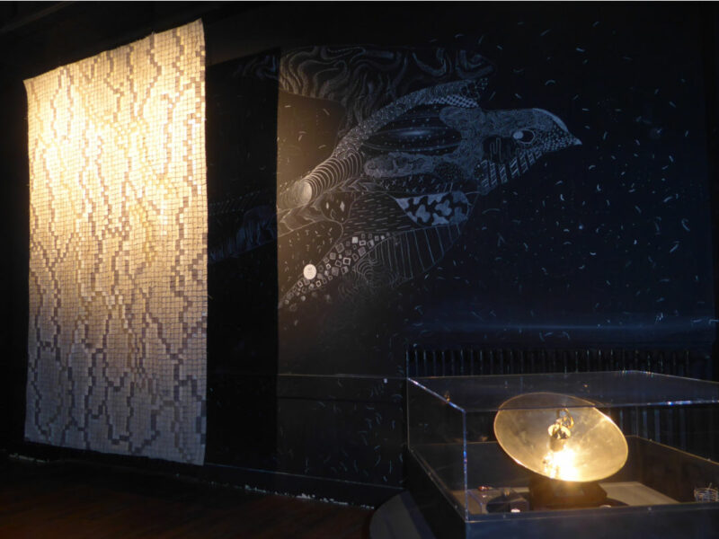 Dimly lit museum exhibit with a tall vertical mosaic wall on the left, a dark mural of a stylized sea creature in the center, and a glass display case containing a glowing brass disk.