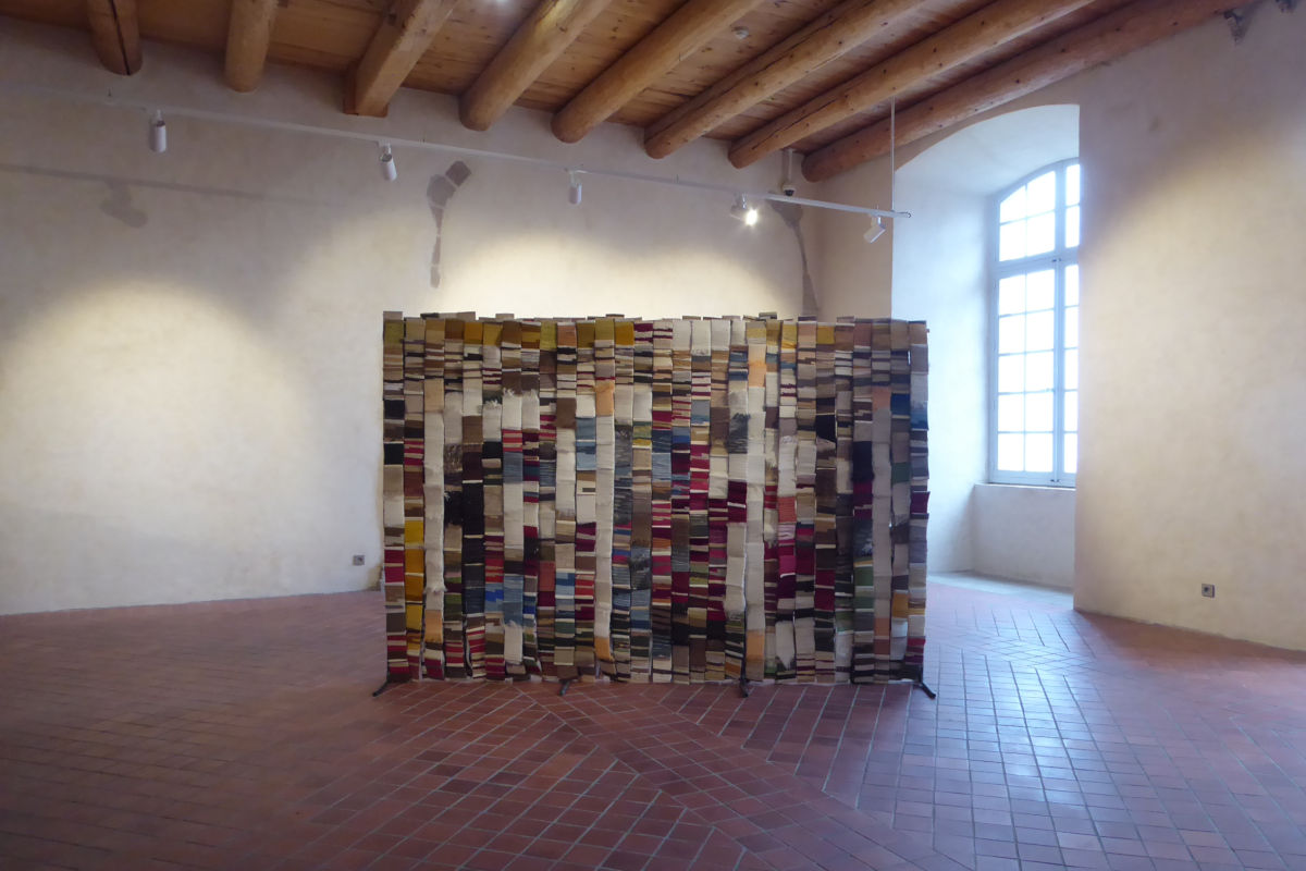 FITE 2020 Regards textiles sur les tapisseries de la Chaise-Dieu ©Thomas Leveugle Installation of stacked books forming a solid screen in a white-walled gallery with a brick floor and wooden ceiling beams; a large window is visible to the right.