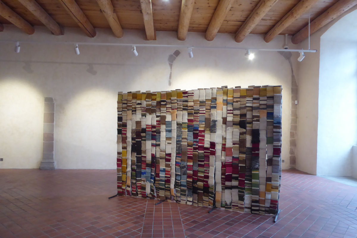FITE 2020 Regards textiles sur les tapisseries de la Chaise-Dieu ©Thomas Leveugle Installation of stacked colored books forming a freestanding screen in a gallery with brick floor and wooden ceiling beams.