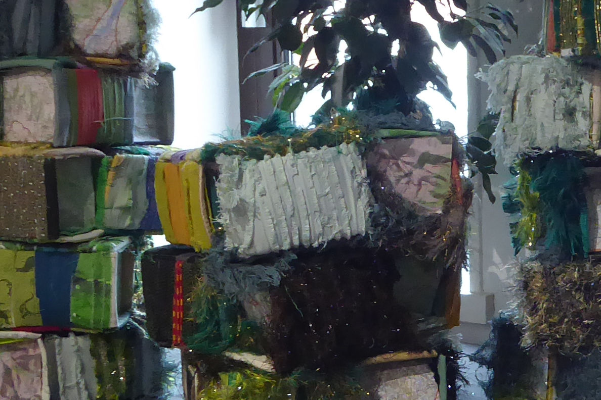 Projet collaboratif - Jardin en hauteur - détail - exposition ©Sabine Cibert Indoor art installation: stacked rectangular blocks wrapped in colorful fabrics and ribbons, with greenery draped over them.