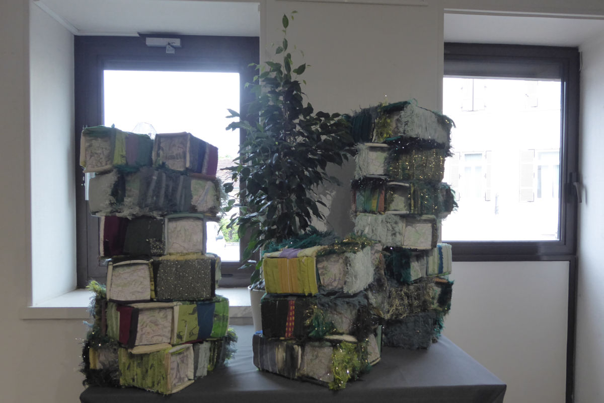 Projet collaboratif - Jardin en hauteur - détail - exposition ©Sabine Cibert Art installation featuring stacked fabric-wrapped blocks on a table near two windows, with a leafy potted plant in the center.