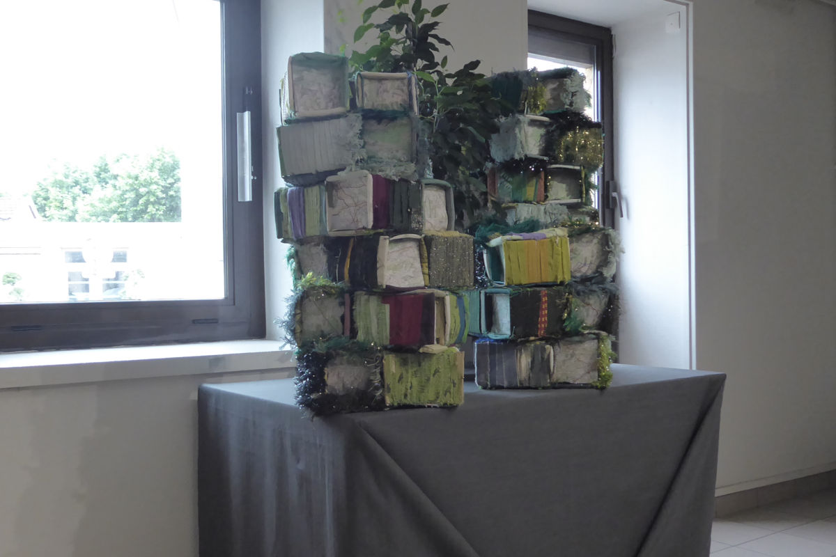 Projet collaboratif - Jardin en hauteur - exposition ©Sabine Cibert Stacked fabric-wrapped rectangular blocks form a tall abstract sculpture on a gray-covered pedestal by a windowed wall.