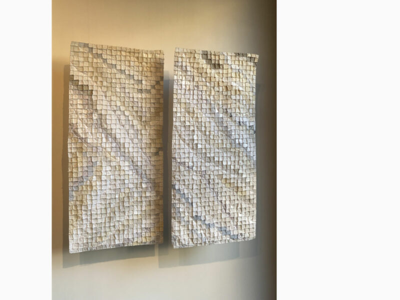 Two vertical mosaic panels made of small square tiles in beige and cream tones mounted on a wall with subtle shadows below.