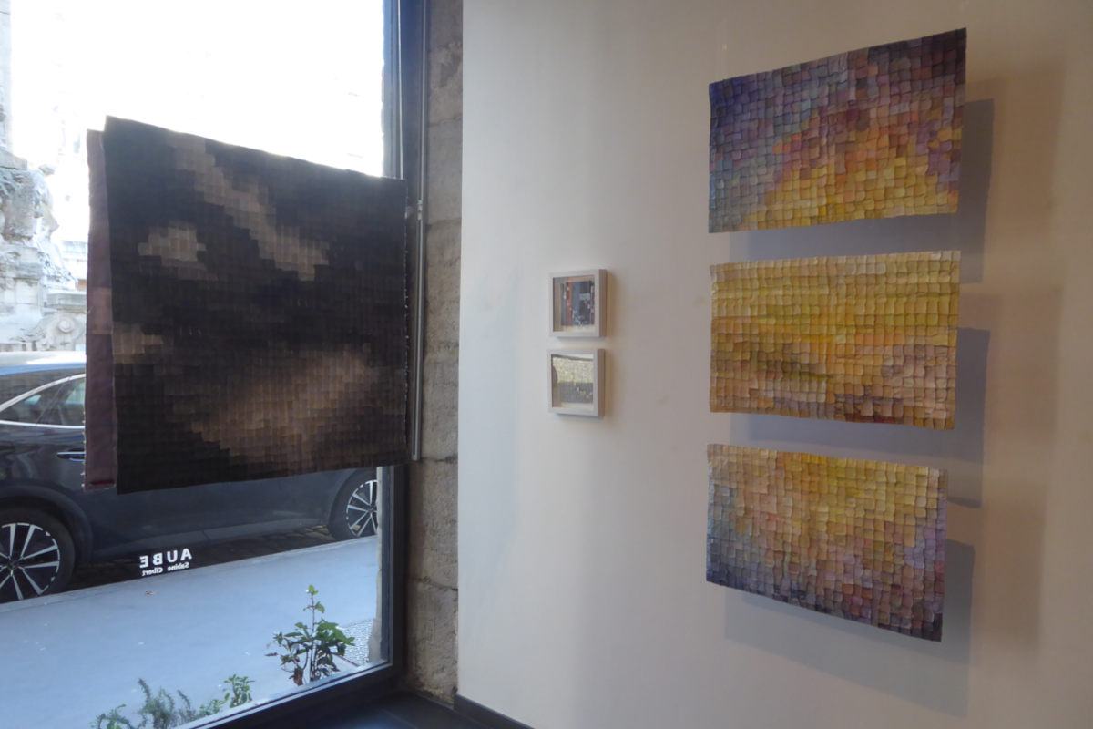 Art gallery interior with three colorful mosaic panels on the right and a large dark square piece near a window on the left, street view outside.