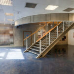 Art gallery interior with a curved metal staircase, wooden handrail, and blue floor, facing quilted wall art on the left.
