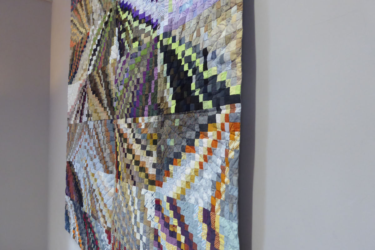 Sabine Cibert Instants 1 détail Exposition Mauriac ©Sabine Cibert_220406_756TB-r Colorful patchwork quilt hanging on a gray wall, made of many small square patches in varied hues.