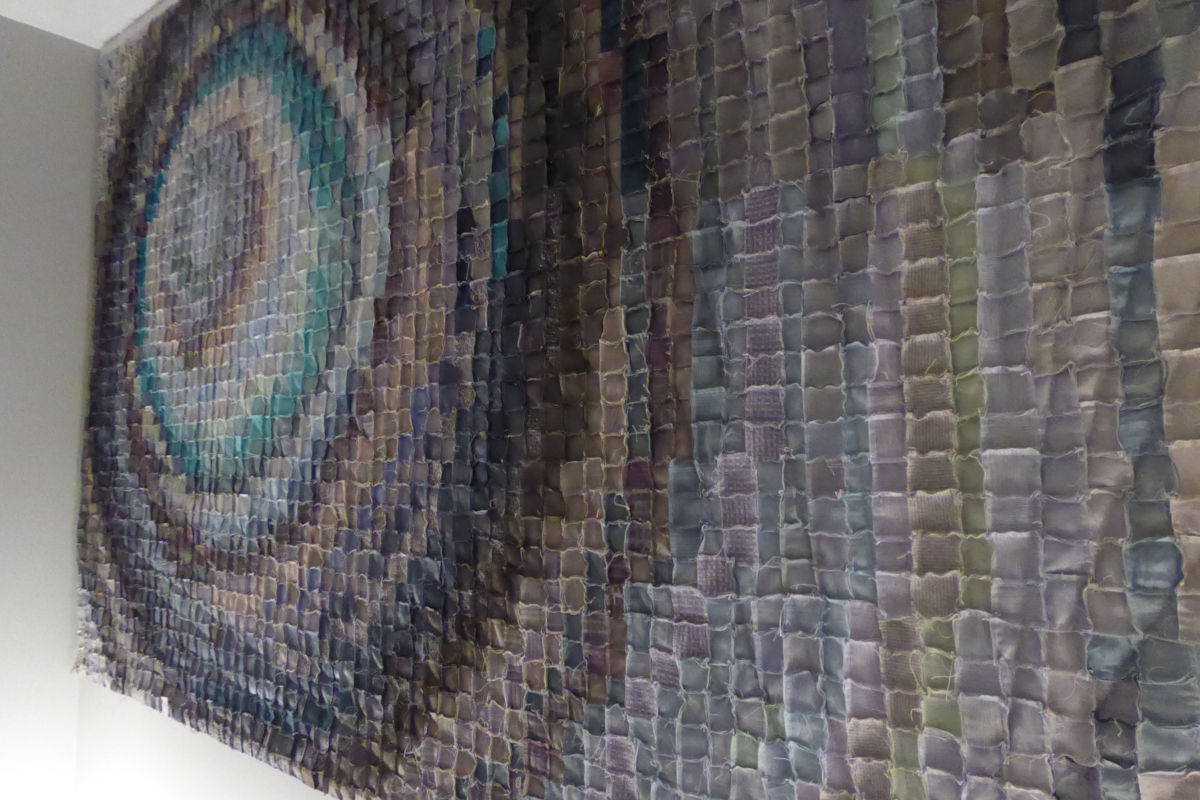 Sabine Cibert Interferences detail In situ ©Sabine Cibert Colorful quilted wall tapestry made of small square fabric patches arranged in a circular pattern on the left, extending across the wall.