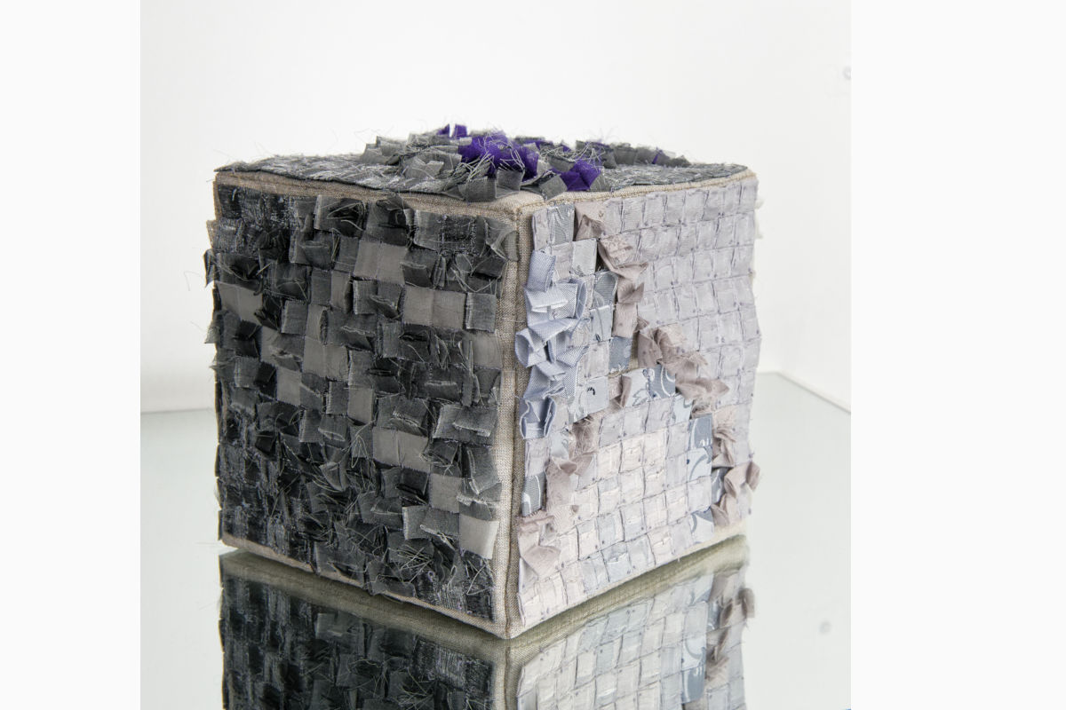 Sabine Cibert- L ira del cielo ©Pascal Muradian Textured fabric cube sculpture made from small square patches in grayscale with a purple accent on top, reflecting on a glass surface