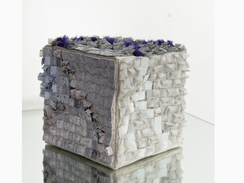 A textured cube sculpture made from small fabric squares in gray, lavender, and white on a reflective base.