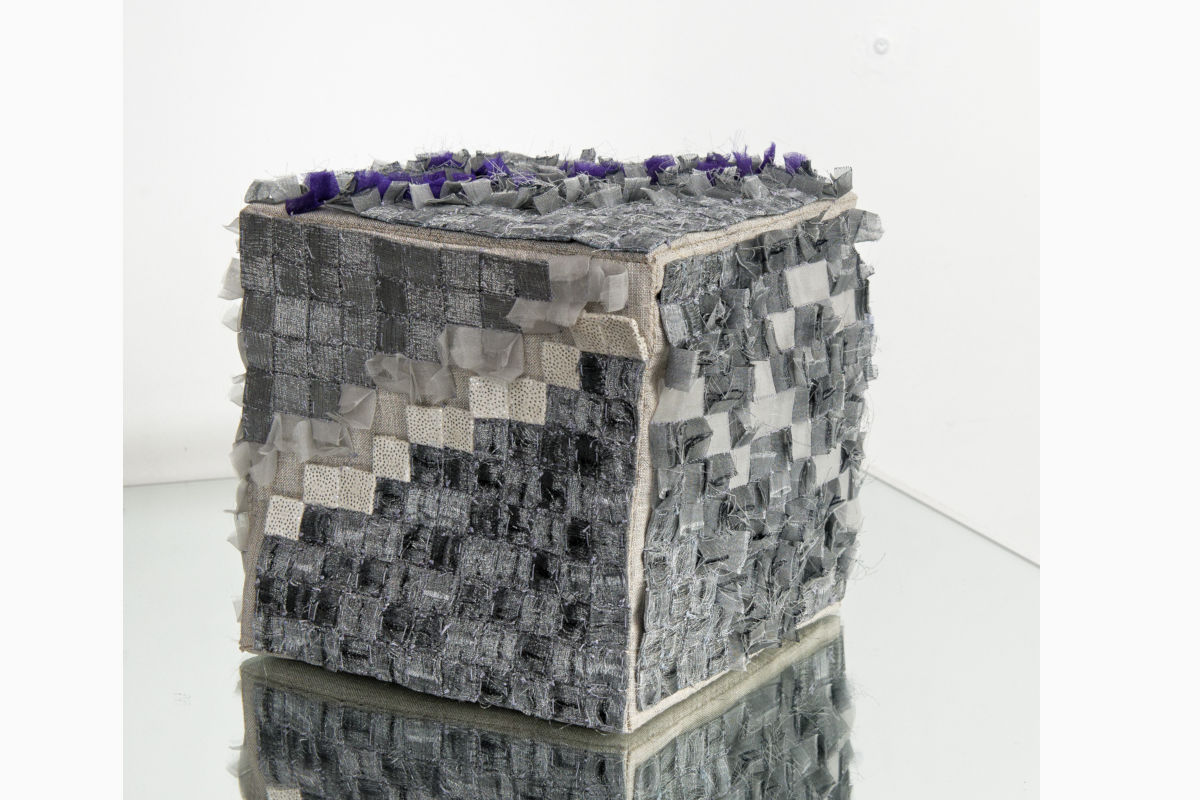 Sabine Cibert- L ira del cielo ©Pascal Muradian Patchwork fabric cube sculpture made of small gray/black squares with purple tips on top, resting on a reflective surface.