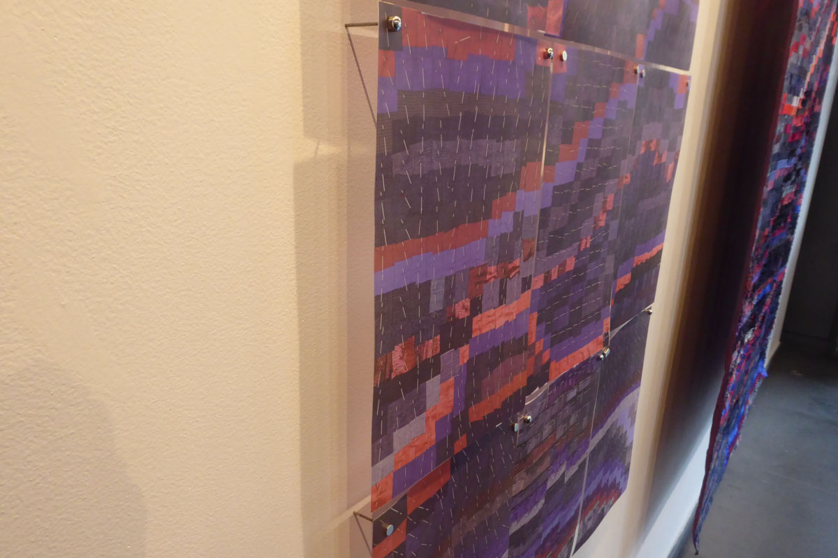 Sabine Cibert - Magma et son archétype détail ©Thomas Leveugle Abstract textile art piece with purple, red, and blue geometric blocks mounted on a beige wall using metal standoffs and clear panels; visible stitching lines across the fabric.