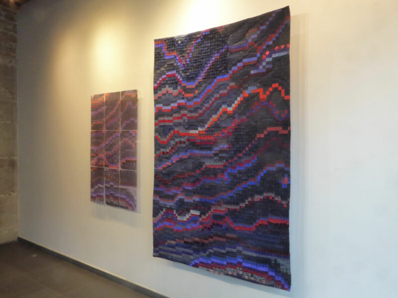 Abstract textile wall hanging with wavy bands of purple, red, and blue in a gallery setting with a smaller matching piece nearby.