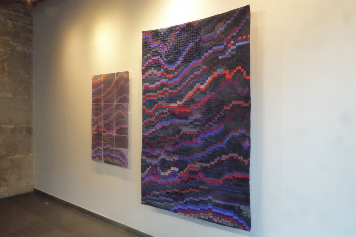 Abstract textile wall hanging with wavy bands of purple, red, and blue in a gallery setting with a smaller matching piece nearby.