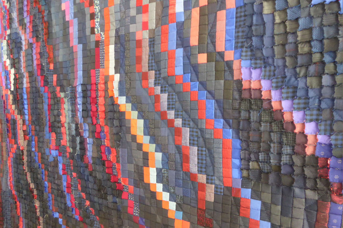 Sabine Cibert - Magma 2 détail ©Sabine Cibert Colorful quilted wall artwork made of small fabric squares in red, blue, purple and gray, arranged in wavy diagonal stripes.