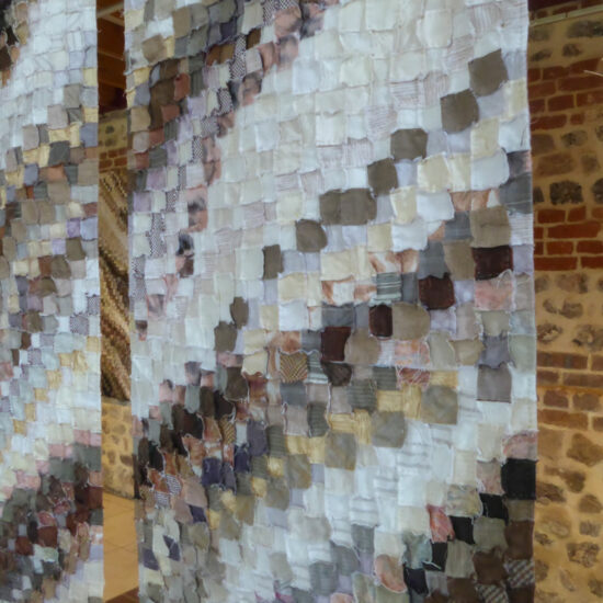 Colorful fabric square panels hanging in long curtains, creating a textured installation in a gallery space on stone walls.