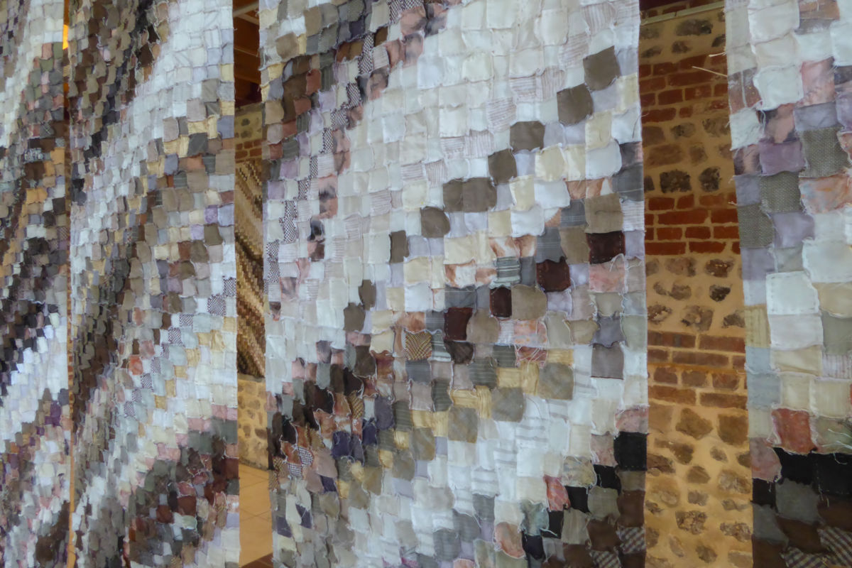 Colorful fabric square panels hanging in long curtains, creating a textured installation in a gallery space on stone walls.