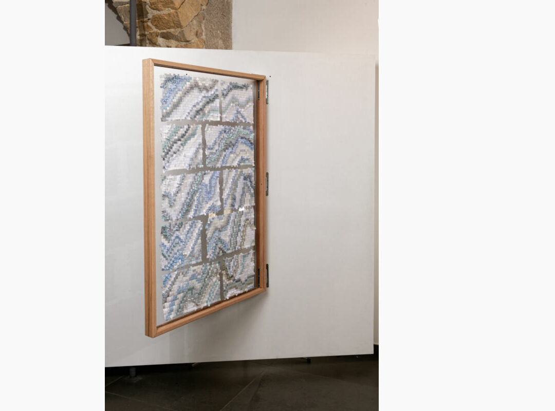 Sabine Cibert Sikuliaq face A de biais ©Pascal Muradian Wood-framed panel mounted on a white wall, filled with a mosaic of small blue and gray tiles forming wavy patterns.