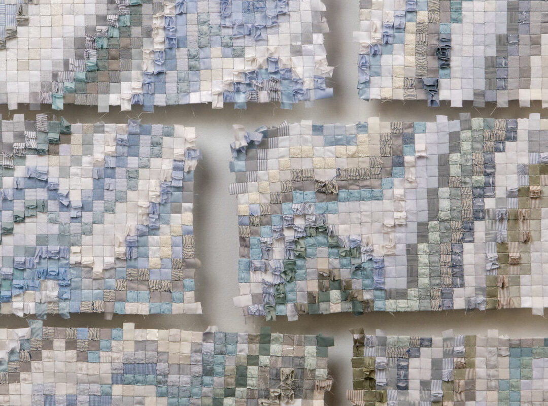 Sabine Cibert Sikuliaq face A détail ©Pascal Muradian Patchwork textile panels arranged in a grid, featuring pastel blue, beige, and gray fabrics forming a quilt-like wall installation in a gallery.