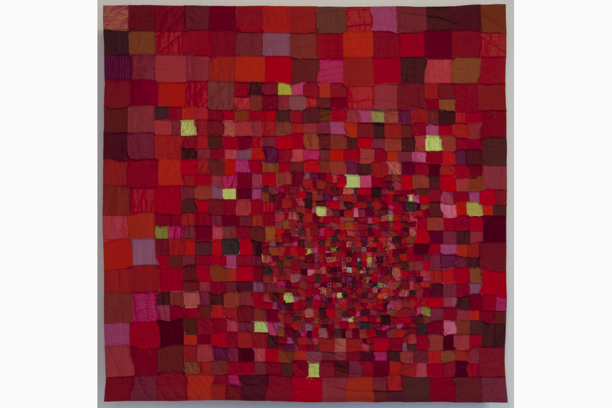 Sabine Cibert Throb 2 ©Philippe-Liev Pourcelot Patchwork quilt of red, pink, and orange squares arranged in a spiral toward the center, displayed on a wall.