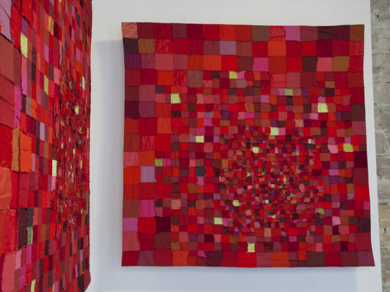 Patchwork quilt of red, pink, and burgundy squares hung on a white wall in a gallery. A second quilt is reflected on the left wall; both are red-toned compositions.