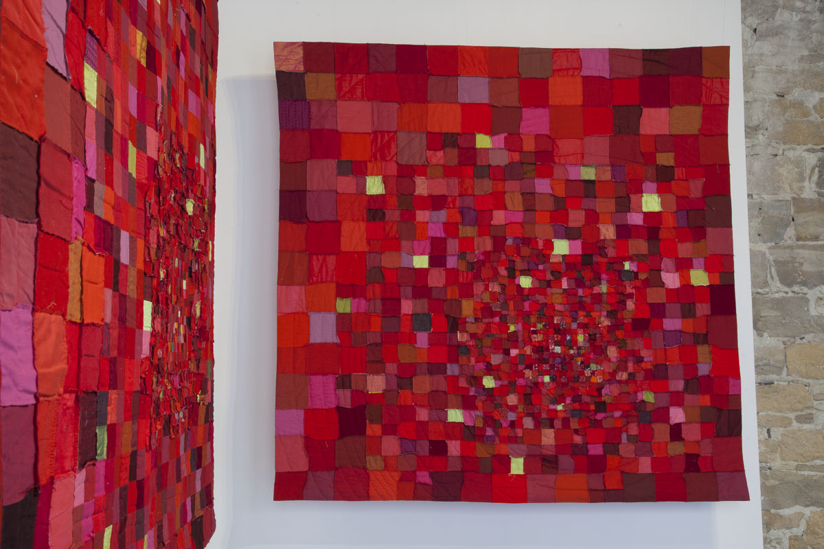 Patchwork quilt of red, pink, and burgundy squares hung on a white wall in a gallery. A second quilt is reflected on the left wall; both are red-toned compositions.