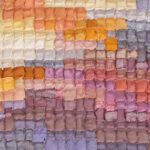 Patchwork quilt made of small pastel fabric squares in warm yellows, pinks, and purples fading across the piece.