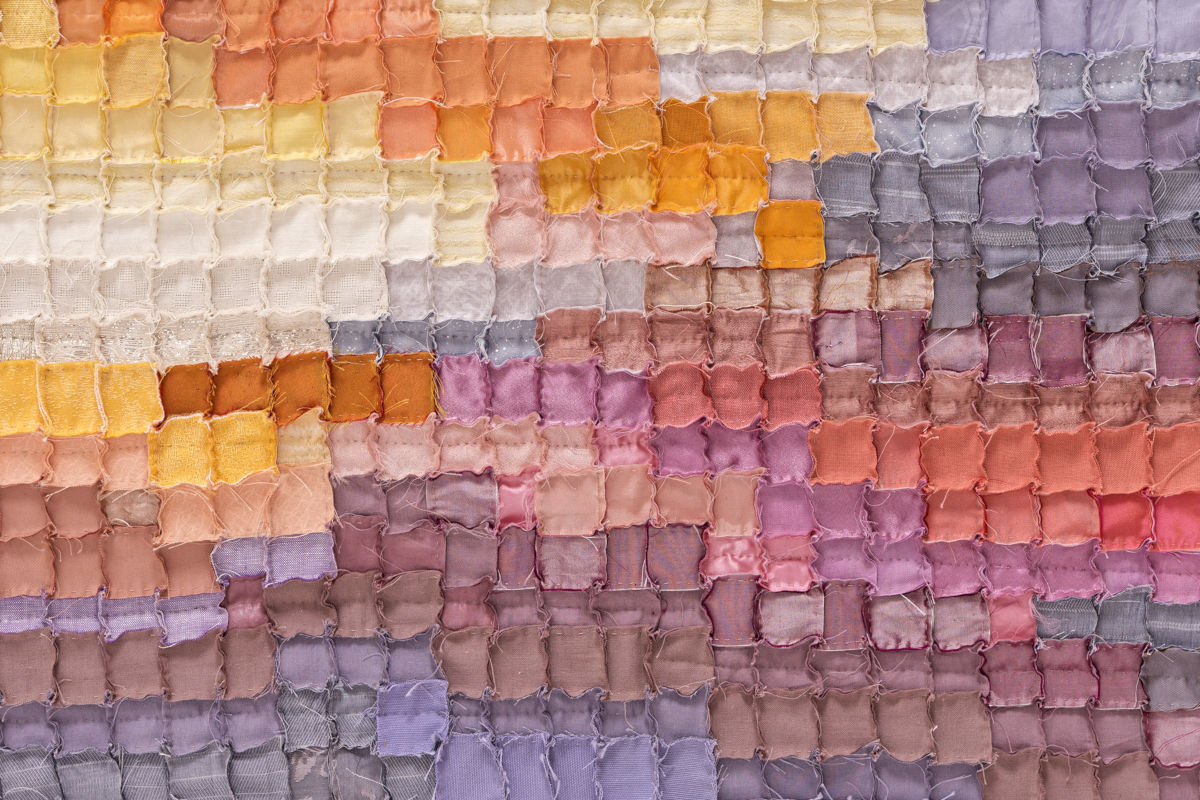 Patchwork quilt made of small pastel fabric squares in warm yellows, pinks, and purples fading across the piece.