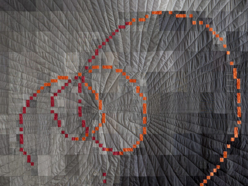 Patchwork quilt featuring a concentric spiral of orange and red squares on gray-toned fabric.
