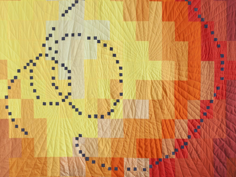 Quilted wall hanging with a large blue-dotted spiral pattern over a yellow-to-orange gradient patchwork quilt.