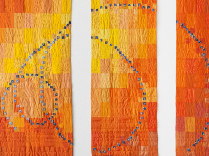 Three vertical quilt panels in orange and yellow squares with a blue dotted spiral curve overlaid as a textile artwork.