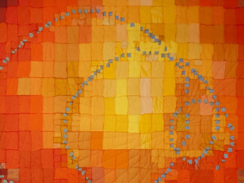 Abstract quilt with orange and yellow patchwork forming a large spiral outlined by small blue squares on a warm-toned background.