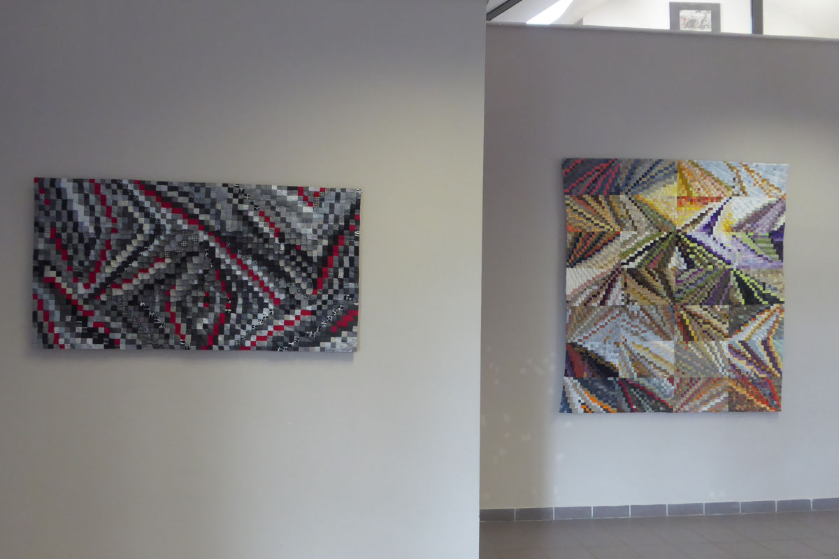 Two abstract mosaic paintings on light gray gallery walls; left piece uses grayscale tiles with red accents in a wavy, zigzag pattern, right piece uses multicolored geometric shards arranged in angular segments.