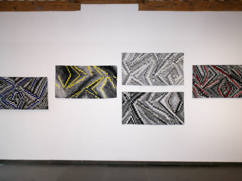 Five rectangular abstract artworks with pixel-like zigzag patterns arranged along a white wall in a gallery. Colors include blue, yellow, and red accents on grayscale backgrounds.